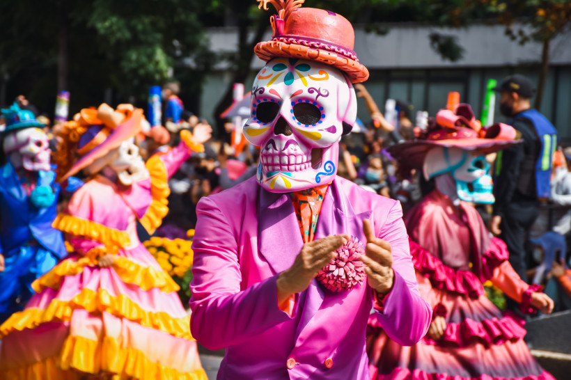 Carnaval in Mexiko Person in einem leuchtend pinken Anzug mit kunstvoll bemalter Totenkopfmaske nimmt an einer Día de los Muertos Parade teil, umgeben von anderen verkleideten Teilnehmern in bunten Kostümen