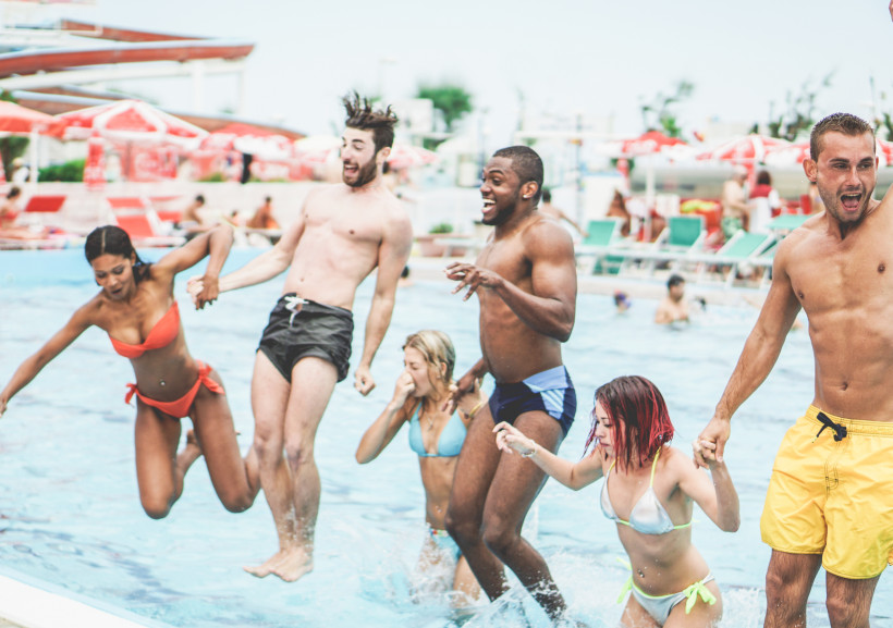 Waterpark-Party am Pool mit springender Gruppe im Wasserbecken. Gruppe junger Erwachsener springt im Freibad in ein Becken, Wasserspritzer im Vordergrund.