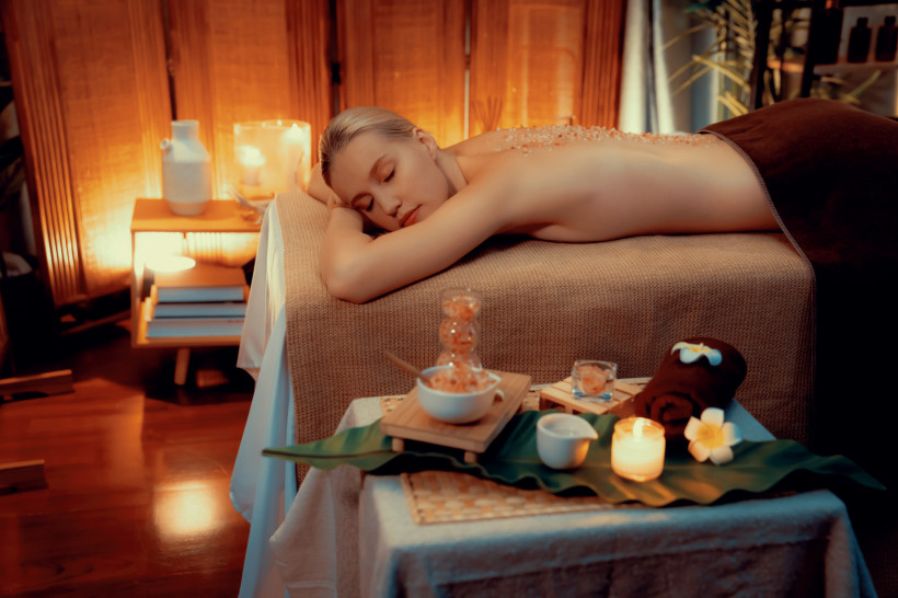 Thailand Pattaya Frau liegt entspannt mit geschlossenen Augen auf einem Massagebett in warm beleuchtetem Spa-Raum. Auf ihrem Rücken liegen grobe Salzsteine, im Vordergrund stehen Kerzen, Pflegeprodukte und eine Blüte auf einem Tisch