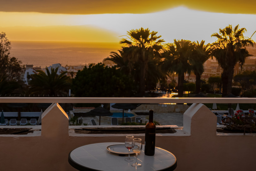 Adeje: Terrasse mit Tisch und Weinflasche vor Palmen und Meer bei Sonnenuntergang Sonnenuntergang über Adeje mit Blick auf Meer, Palmen und Terrasse mit Tisch, Weinflasche und zwei Gläsern