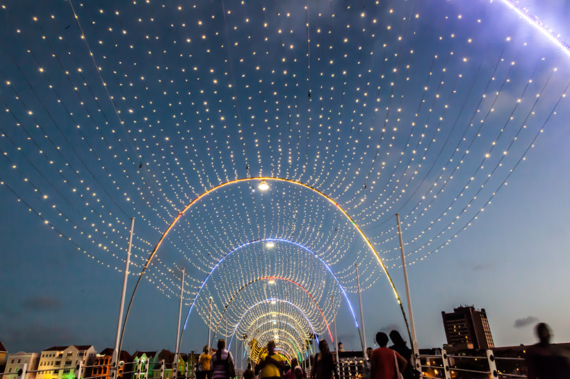 Curaçao mma-Brücke in Willemstad, Curaçao, am AbeDie berühmte Königin-End. Über der Brücke spannen sich mehrere Bögen aus leuchtenden Lichterketten. Menschen spazieren unter dem funkelnden Lichterhimmel hindurch. Am Rand sind bunte Häuserfassaden zu erkennen