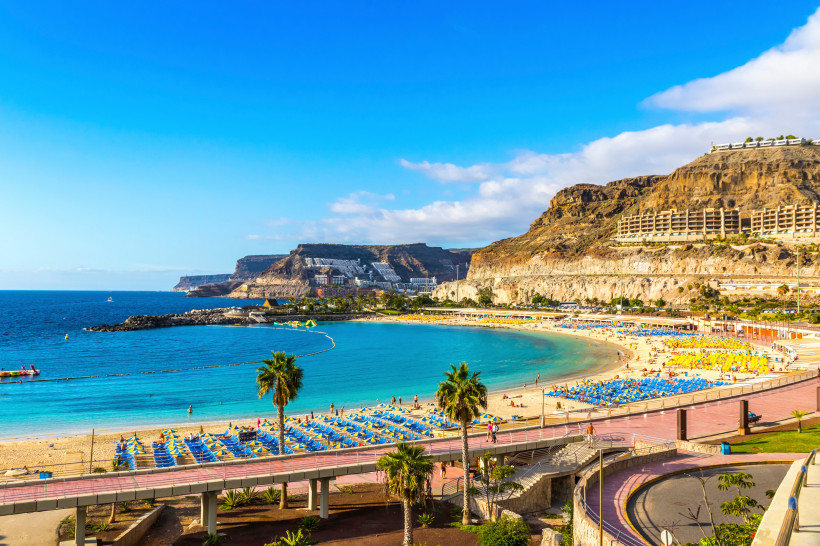 Traumstrand von Amadores – Sonnenurlaub auf Gran Canaria Panoramablick auf den Strand von Amadores auf Gran Canaria mit türkisblauem Meer, goldgelbem Sand, Sonnenschirmen und Sonnenliegen. Im Hintergrund erheben sich Felsen mit Hotels und Promenade unter blauem Himmel.