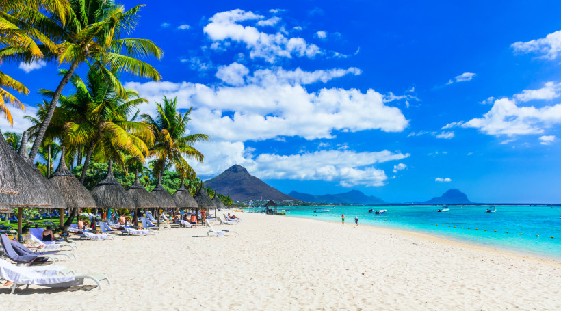 Weißer Sandstrand mit Palmen und Strohdach-Liegen am Meer in Flic en Flac auf Mauritius, mit dem Berg Le Morne im Hintergrund
