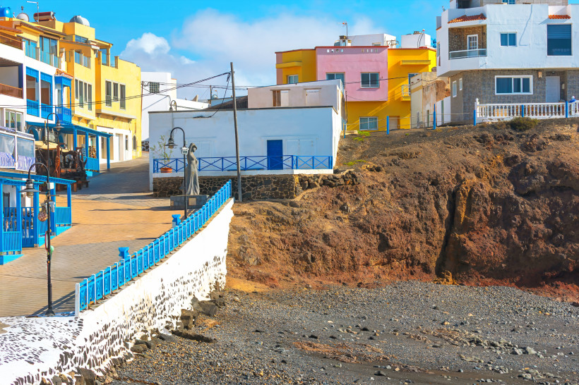 Küstenpromenade in El Cotillo mit bunten Häusern und blauem Geländer oberhalb einer felsigen Bucht