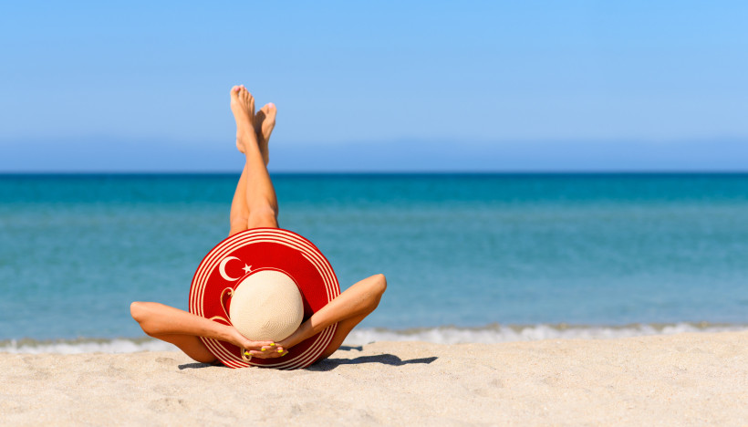Entspannte Urlaubsszene am Strand mit Person im Sand, rotem Sonnenhut und Blick auf das türkisblaue Meer