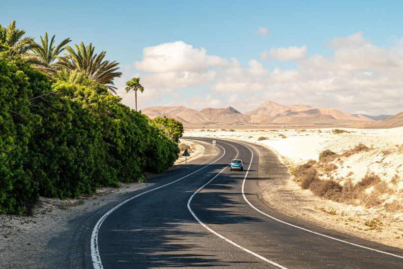 Küstenstraße durch Wüstenlandschaft mit Palmen auf Fuerteventura Auto fährt auf einer Landstraße durch eine trockene, bergige Wüstenlandschaft mit Palmen auf einer Seite und Sanddünen auf der anderen unter blauem Himmel mit Wolken