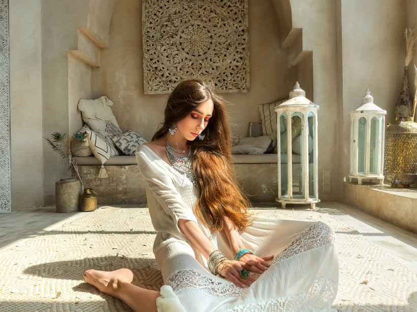 Ägypten - Marsa Alam Frau in weißem Kleid sitzt entspannt im Schneidersitz in einem orientalischen Raum mit Laternen, Kissen und kunstvollen Wänden. Sonnenlicht fällt auf den hellen Teppich.