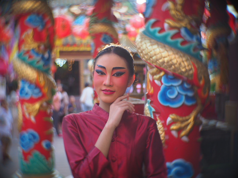 Thailand - Phuket Porträt einer Frau in roter traditioneller Kleidung und dramatischem Bühnen-Make-up. Sie steht vor kunstvoll verzierten, roten Tempelsäulen mit goldenen Drachenmotiven.