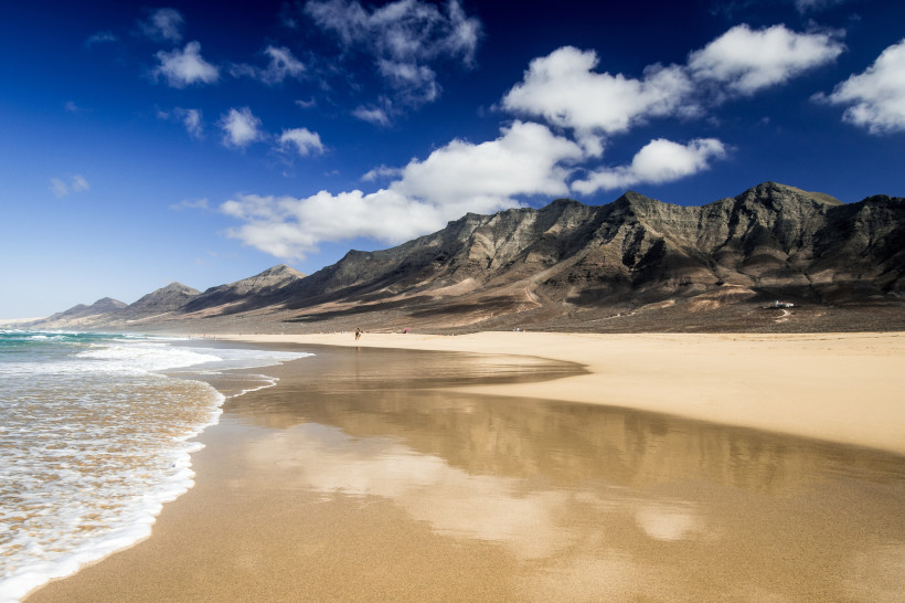 Fuerteventura Weitläufiger, menschenleerer Sandstrand mit sanften Wellen. Im Hintergrund eine zerklüftete Gebirgskette unter blauem Himmel mit vereinzelten Wolken