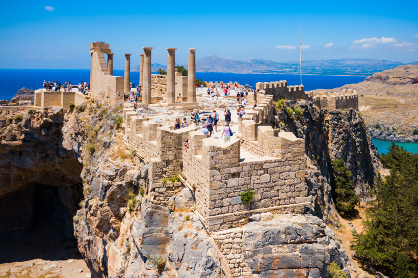 Das Bild zeigt die Akropolis von Lindos auf der griechischen Insel Rhodos, eine gut erhaltene antike Burganlage auf einem steilen Felsplateau.   Die Festung besteht aus massiven, hellen Steinmauern mit Zinnen, Treppen und historischen Säulen, von denen ei