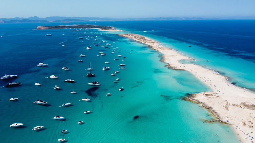 Formentera Luftaufnahme einer langen, schmalen Sandbank in Formentera, Spanien. Zahlreiche weiße Boote und Yachten ankern im türkisblauen, klaren Wasser auf beiden Seiten der Landzunge. Die Szenerie erstreckt sich bis zum Horizont, wo sanfte Hügel und das offene Mee