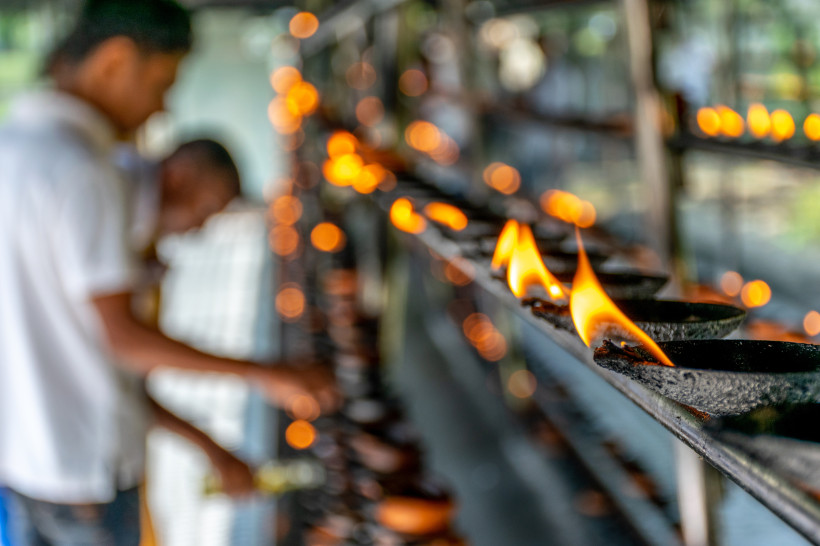 Reihe kleiner Ton-Öllampen mit offenen Flammen auf einer Metallablage im Tempel. Im Hintergrund sind unscharf zwei Menschen zu sehen, die vermutlich weitere Lampen entzünden. Das Bild vermittelt eine ruhige, spirituelle Atmosphäre.