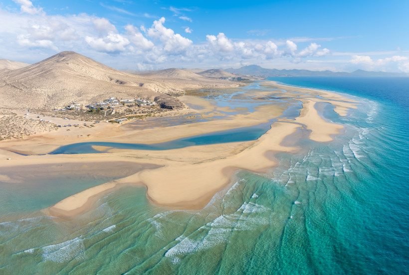 Weite Lagunenlandschaft an der Küste, Playa de Sotavento – Fuerteventura Luftaufnahme einer weiten Küstenlandschaft mit türkisfarbenem Meer, Lagunen, Sandbänken, einem kleinen Dorf und trockenen Berghügeln im Hintergrund