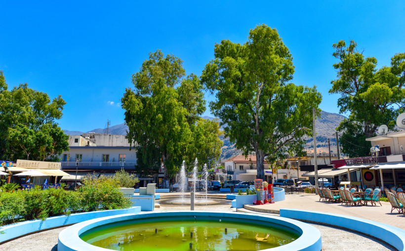 Dorfplatz in Georgioupolis mit Springbrunnen, Bäumen und umliegenden Cafés vor Bergkulisse