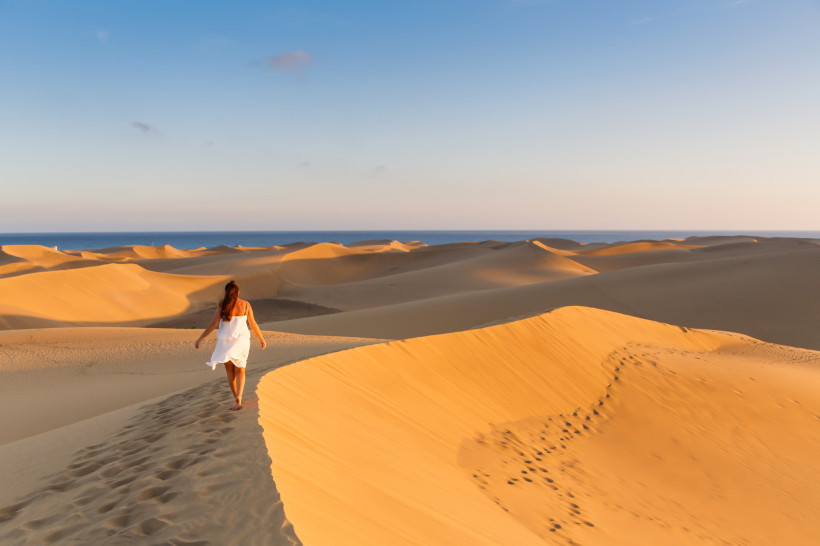 Maspalomas Dünen Gran Canaria Frau in weißem Kleid geht durch die goldgelben Dünen von Maspalomas auf Gran Canaria, im Hintergrund das Meer unter blauem Himmel.