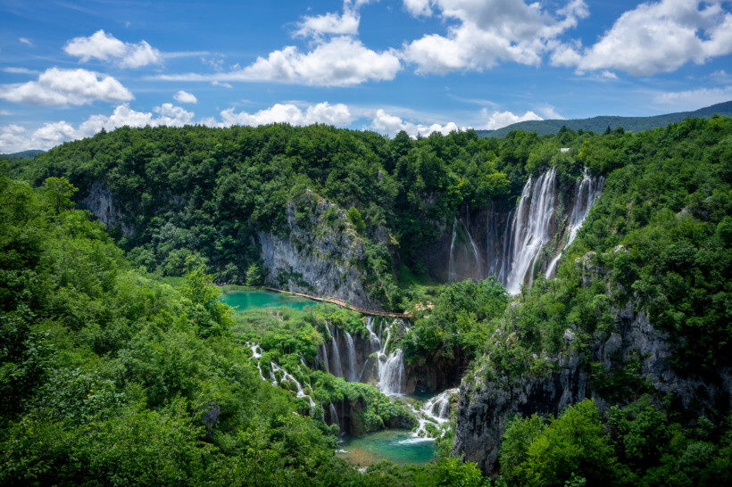 Panoramablick auf die Plitvicer Seen in Kroatien. Mehrere Wasserfälle stürzen von bewaldeten Felswänden in türkisfarbene Seen, die durch Holzstege miteinander verbunden sind. Der Himmel ist blau mit einigen weißen Wolken.