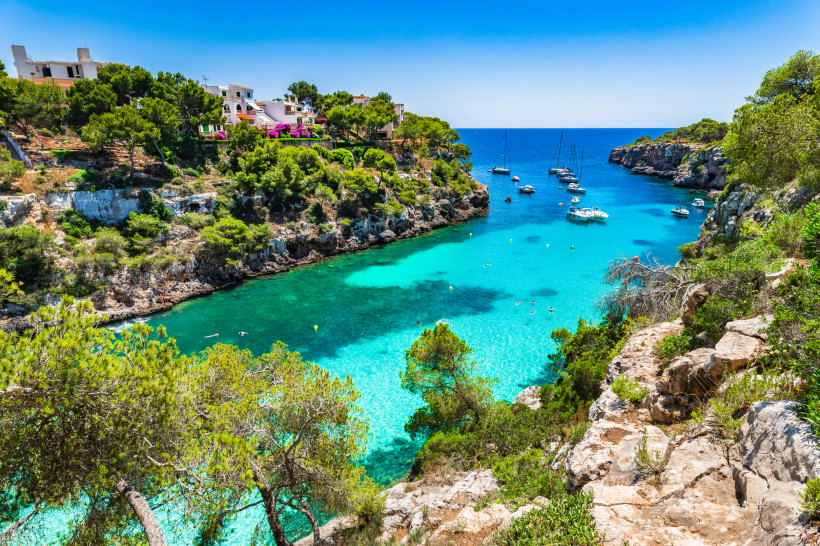 Mallorca - Cala Pi Eine idyllische Bucht auf Mallorca mit klarem, türkisblauem Wasser, gesäumt von grünen Pinien und steilen Klippen. Segelboote und Yachten schwimmen im Wasser, während sich einige Menschen in der Bucht entspannen. Rechts im Bild windet sich ein schmaler Pf