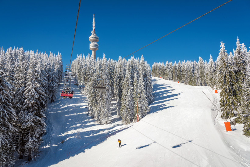 Bulgarien Winterlandschaft im Skigebiet Pamporovo in Bulgarien. Ein Sessellift fährt durch verschneite Tannenwälder hinauf zu den Pisten, während ein einzelner Skifahrer die weiße Piste hinuntergleitet. Im Hintergrund erhebt sich der markante Fernsehturm Snezhanka