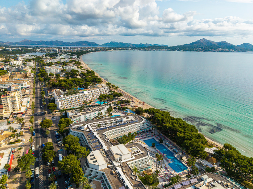 Luftaufnahme der Playa de Alcudia mit breitem Sandstrand, Hotelanlagen, Strandpromenade und flach abfallendem Wasser entlang der Bucht