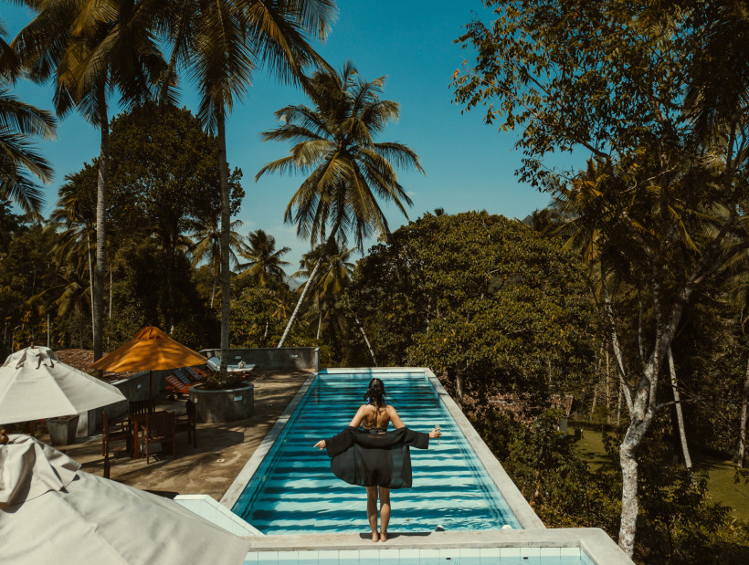 Sri Lanka Eine Frau in schwarzer Badebekleidung steht am Rand eines Infinity-Pools, der von Palmen und dichtem tropischem Grün umgeben ist. Sie schaut in die Ferne, die Arme leicht ausgebreitet. Im Hintergrund stehen Sonnenschirme und Holzmöbel auf der Terrasse. Di