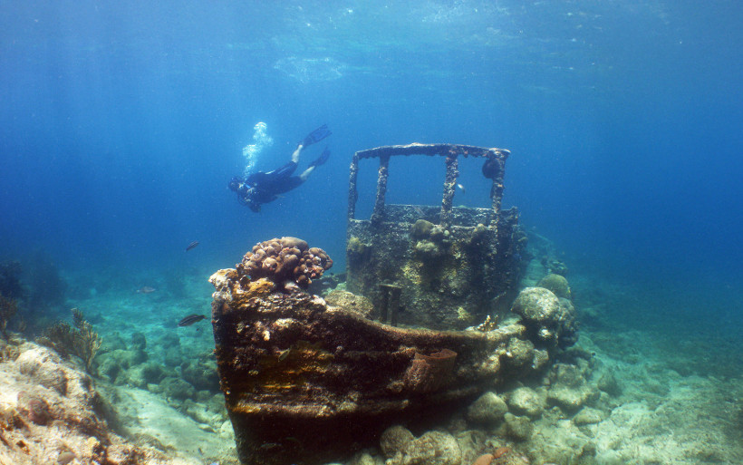 Curaçao Taucher erkundet ein versunkenes Schiffswrack unter Wasser. Das Wrack ist teilweise mit Korallen bewachsen und liegt auf einem felsigen Meeresboden. Im klaren blauen Wasser sind einige Fische zu sehen, während Sonnenlicht durch die Wasseroberfläche dringt