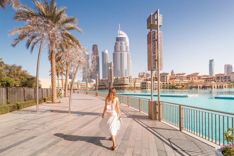 Dubai Urlaub Eine Frau in weißem Kleid spaziert an einer sonnigen Uferpromenade in Dubai entlang. Im Hintergrund ragen moderne Wolkenkratzer in den blauen Himmel, Palmen säumen den Weg, und das Wasser einer künstlichen Lagune glänzt türkis im Sonnenlicht.