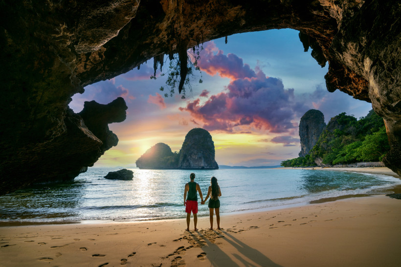 Thailand - Krabi Das Bild zeigt ein Paar, das Hand in Hand am Rand eines tropischen Sandstrandes steht. Sie befinden sich in einer natürlichen Felsbucht, eingerahmt von dunklem, zerklüftetem Gestein. Vor ihnen liegt das ruhige Meer, in dem sich das warme Licht eines farbe