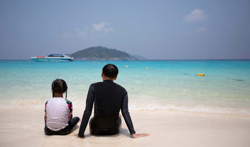 Mädchen und Junge sitzen am Strand im Sand und blicken auf das türkisfarbene Meer mit Insel und Boot im Hintergrund.