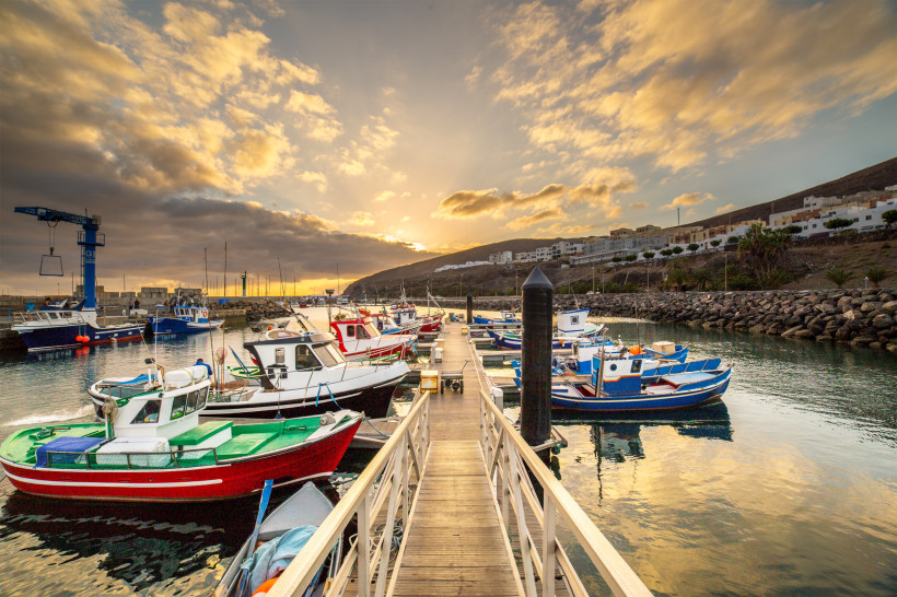 Bunte Fischerboote im Hafen von Gran Tarajal auf Fuerteventura bei Sonnenuntergang, mit Blick auf den ruhigen Atlantik und die umliegenden Hügel.