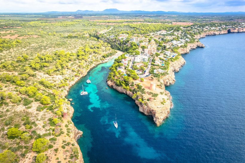 Luftaufnahme der Bucht Cala Pi mit türkisblauem Wasser, Steilküste, Segelbooten und Siedlung