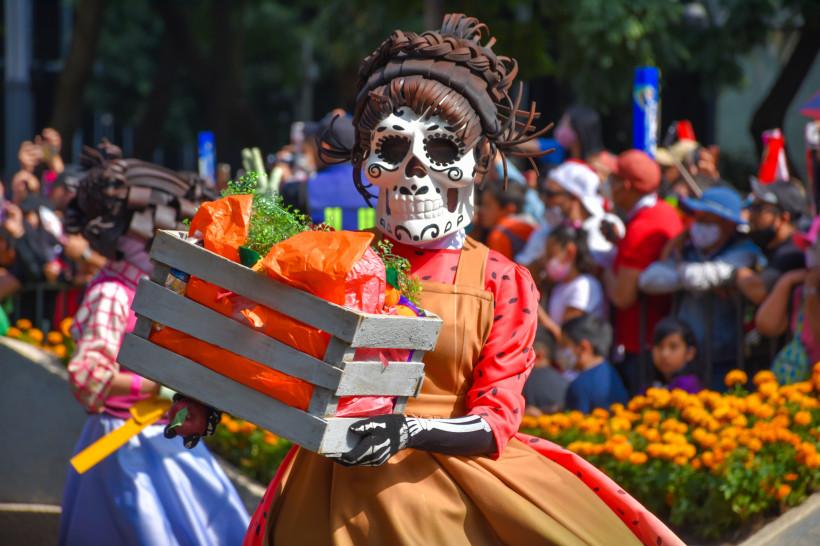 Eine festlich gekleidete Teilnehmerin der Día de los Muertos Parade trägt eine kunstvolle Calavera-Maske und ein traditionelles Kleid in lebendigen Farben. Sie hält eine mit Blumen und bunten Stoffen dekorierte Kiste, während im Hintergrund eine jubelnde 