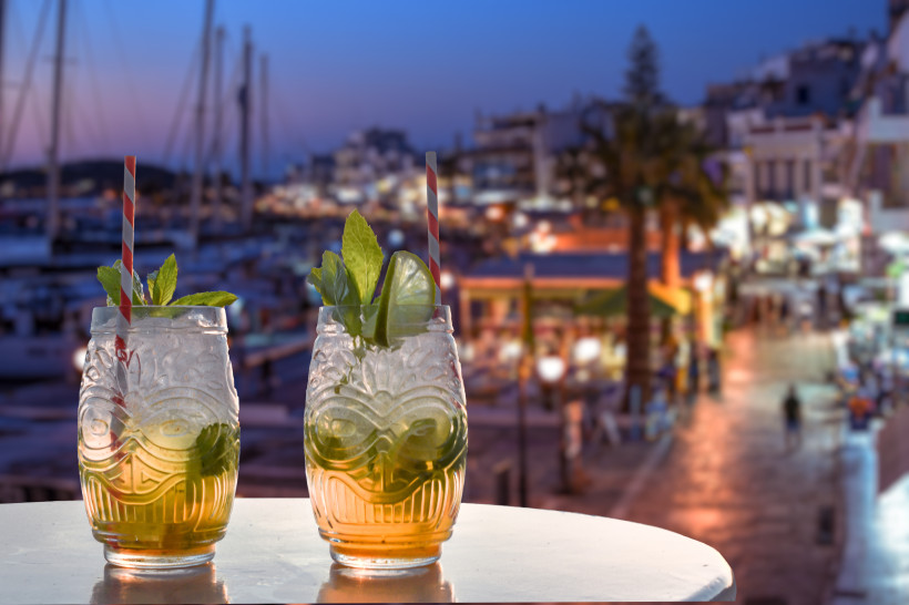 Mallorca Zwei Cocktails in verzierten Gläsern mit Minzblättern, Limettenscheiben und gestreiften Trinkhalmen stehen auf einem Tisch. Im Hintergrund unscharf eine abendliche Hafenpromenade mit Lichtern, Restaurants und Bootsmasten.