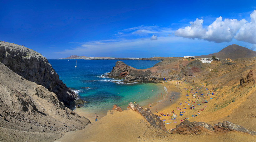 Belebter Sandstrand in einer geschützten Bucht mit Badegästen, umgeben von Felsen, blauem Meer, weißen Häusern am Hang und einer weiten Vulkanlandschaft