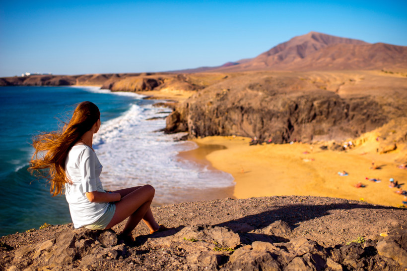 Lanzarote Reisearten – Traumstrände, Naturerlebnisse Frau sitzt auf einer Klippe mit Blick auf eine einsame Bucht auf Lanzarote – ideal für Natur- und Strandurlaub