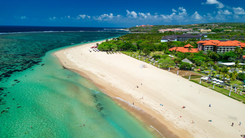 Panoramablick auf einen langen, breiten Sandstrand mit türkisfarbenem Wasser bei Nusa Dua auf Bali. Der Strand ist von einigen wenigen Badegästen besucht und wirkt ruhig und sauber. Im Hintergrund liegen Resorts mit roten Dächern, eingebettet in viel trop