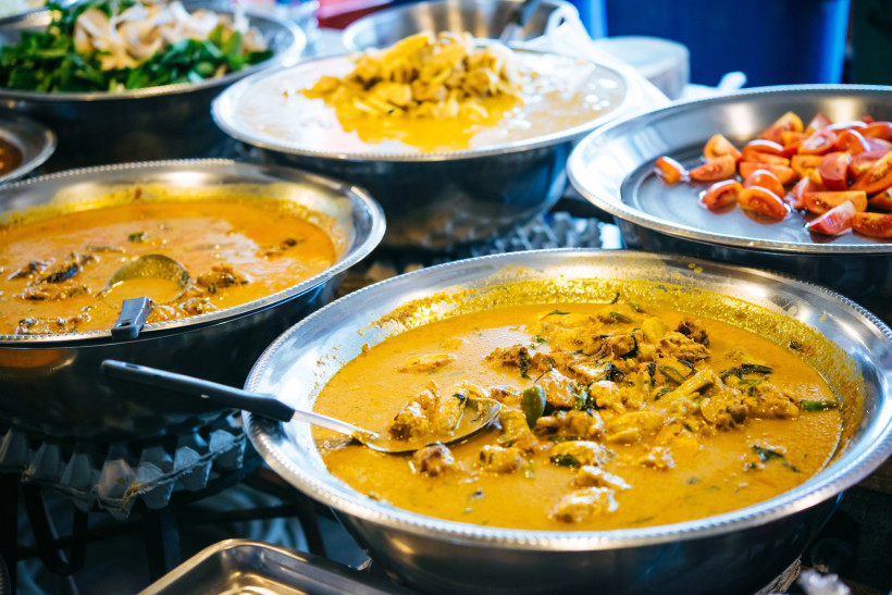 Thailand - Koh Samui  Das Bild zeigt mehrere große silberne Metallschalen, gefüllt mit typischen thailändischen Currys und Zutaten. Im Vordergrund befinden sich zwei gelb-orangene Currys mit Gemüse, Kräutern und Fleischstücken, aus denen große Löffel ragen. In weiteren Schale