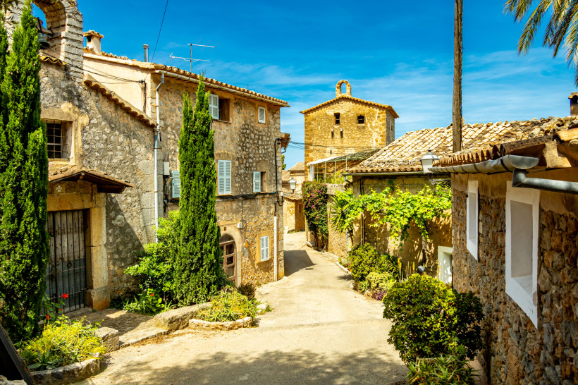 Mallorca - Deià Ein malerisches Dorf auf Mallorca mit traditionellen Steinhäusern, grün gestrichenen Fensterläden und engen Kopfsteinpflastergassen. Die Häuser sind von grünen Pflanzen und bunten Blumen geschmückt. Eine kleine Kirche mit einem Glockenturm erhebt sich im
