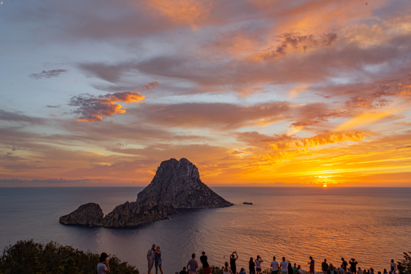 Ibiza - Es Vedrà Menschen beobachten den Sonnenuntergang mit Blick auf die Felseninsel Es Vedrà. Der Himmel ist orange-rot gefärbt, die Sonne versinkt im Meer hinter der markanten Insel.