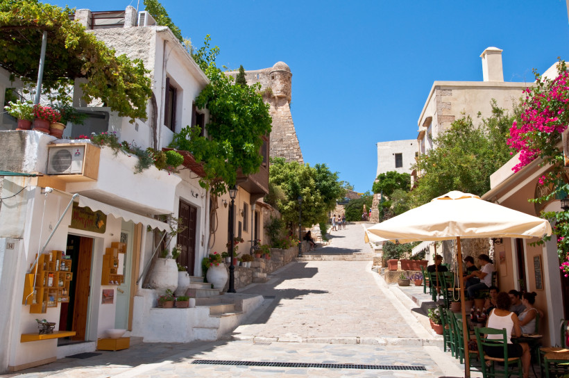Gasse in der Altstadt von Rethymno mit weißen Häusern, Cafétischen und blühenden Bougainvilleen