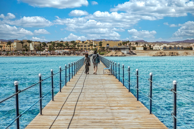Holzsteg über dem türkisblauen Meer in Marsa Alam, zwei Personen spazieren Richtung Strand und Hotels