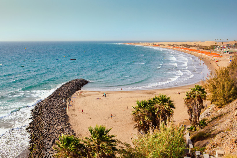 Blick über den Strand von Playa del Inglés mit Wellen, Wellenbrecher aus Steinen und Dünen im Hintergrund