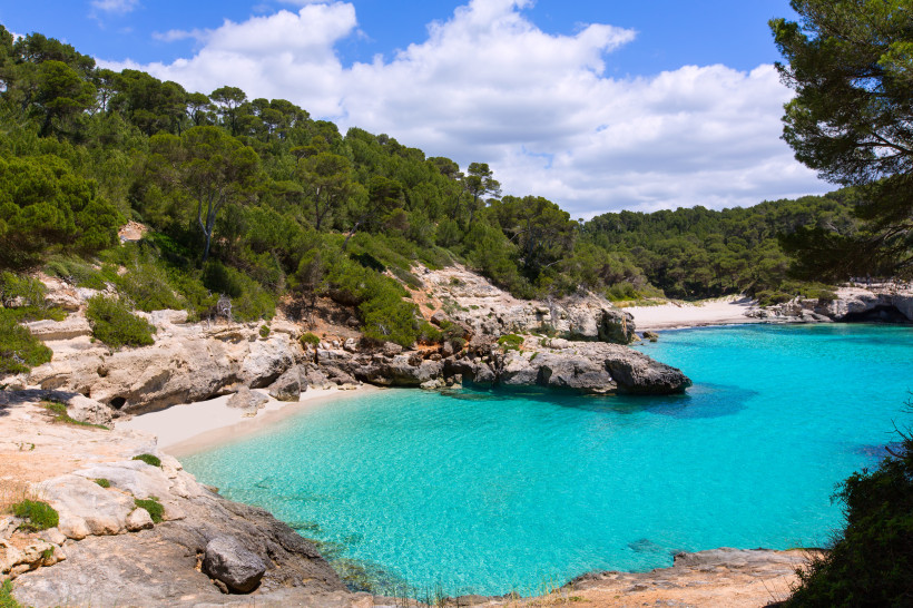 Kleine Badebucht Cala Mitjana und Mitjaneta auf Menorca: klares, türkisfarbenes Wasser, helle Felsen und dichter Pinienwald, die den weißen Sandstrand umrahmen.