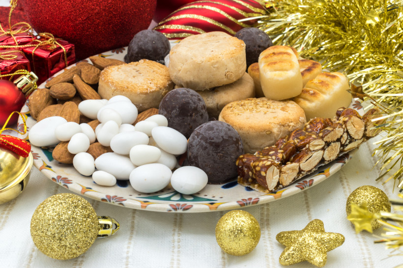 Ein dekorativer Weihnachtsteller mit typisch spanischem Weihnachtsgebäck: Polvorones, Turrón, Marzipanfiguren, kandierten Mandeln und Schokoladenkugeln, umrahmt von goldenen Christbaumkugeln und Weihnachtsdekoration.