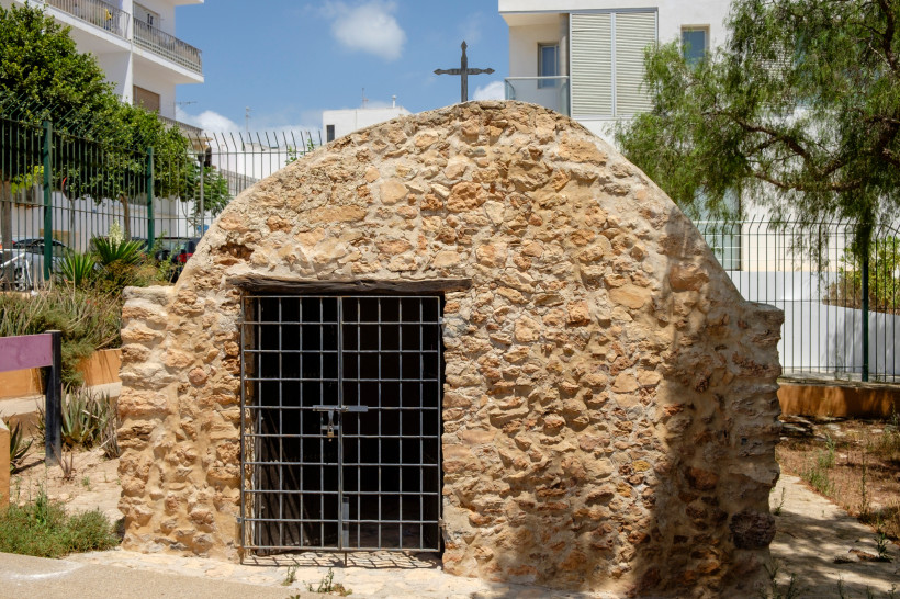 Formentera Kleine Kapelle aus Naturstein mit Gittertür und einem Kreuz auf dem Dach, umgeben von moderner Bebauung und einem Zaun. Der Himmel ist blau mit wenigen Wolken.