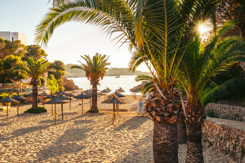 Sandstrand in Cala d’Or mit Palmen, Strohdächern und ruhigem Wasser im warmen Abendlicht