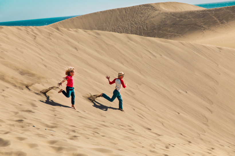 Zwei Kinder laufen eine Sanddüne in den Dunas de Maspalomas nahe Meloneras hinunter, im Hintergrund das Meer.