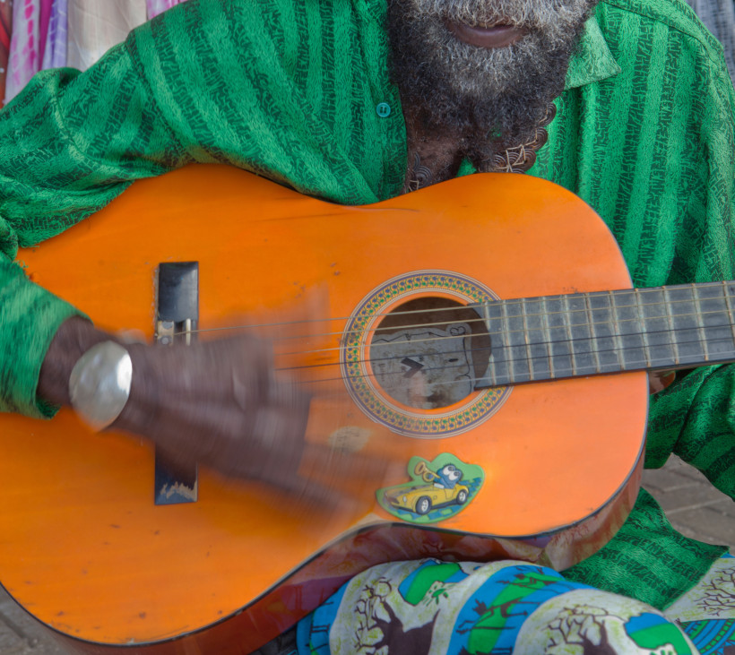 Curaçao Nahaufnahme eines Musikers mit dunkler Haut und Bart, der eine orangefarbene, abgenutzte Gitarre spielt. Die Saiten sind in Bewegung, wodurch die Hand verschwommen wirkt. Der Musiker trägt ein grünes Hemd mit Muster und einen auffälligen silbernen Armreif