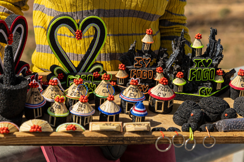 Handgemachte Souvenirs aus Fogo, Kapverden, auf einem Holztisch ausgestellt. Zu sehen sind Miniaturhütten mit Strohdächern, Lavasteinfiguren mit der Aufschrift 'I ❤️ Fogo' und Schlüsselanhänger. Im Hintergrund trägt eine Person einen gelb-grau gestreiften