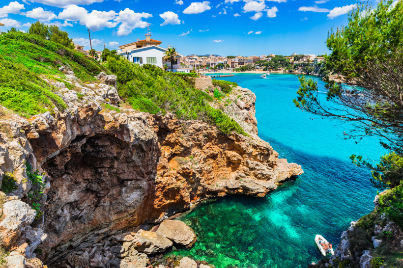 Mallorca - Porto Cristo Panoramaaufnahme von Porto Cristo mit dem türkisfarbenen Hafenbecken und der Stadt dahinter. Weiße und sandfarbene Häuser reihen sich dicht aneinander, viele mit Flachdächern oder Dachterrassen. Im Hafen liegen zahlreiche Boote und Yachten. Rechts führt e