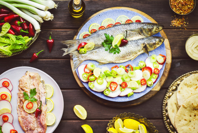 Ägypten - Hurghada Zwei ganze gesalzene Fische auf einem blauen Teller, umgeben von Limettenscheiben, Chili, Frühlingszwiebeln und Radieschen. Daneben verschiedene Beilagen wie Fladenbrot, Zitronenschnitze und Gewürze, auf einem dunklen Holztisch angerichtet.